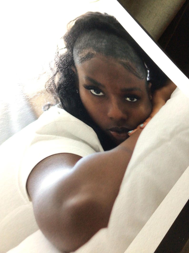 May include: A person with dark hair styled in a partial shave on the side, wearing a plain white t-shirt. The individual is resting their head on a white pillow, looking directly toward the camera with a neutral expression and dark eyeliner.