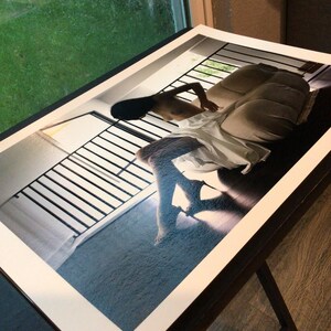 May include: A rectangular photographic print featuring a high-contrast scene of a figure reclining on a beige sofa near a window with black railings. The figure wears a white garment, and the composition emphasizes deep shadows and bright light.