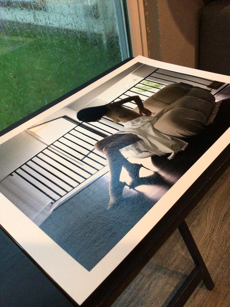 May include: A rectangular photographic print featuring a high-contrast scene of a figure reclining on a beige sofa near a window with black railings. The figure wears a white garment, and the composition emphasizes deep shadows and bright light.