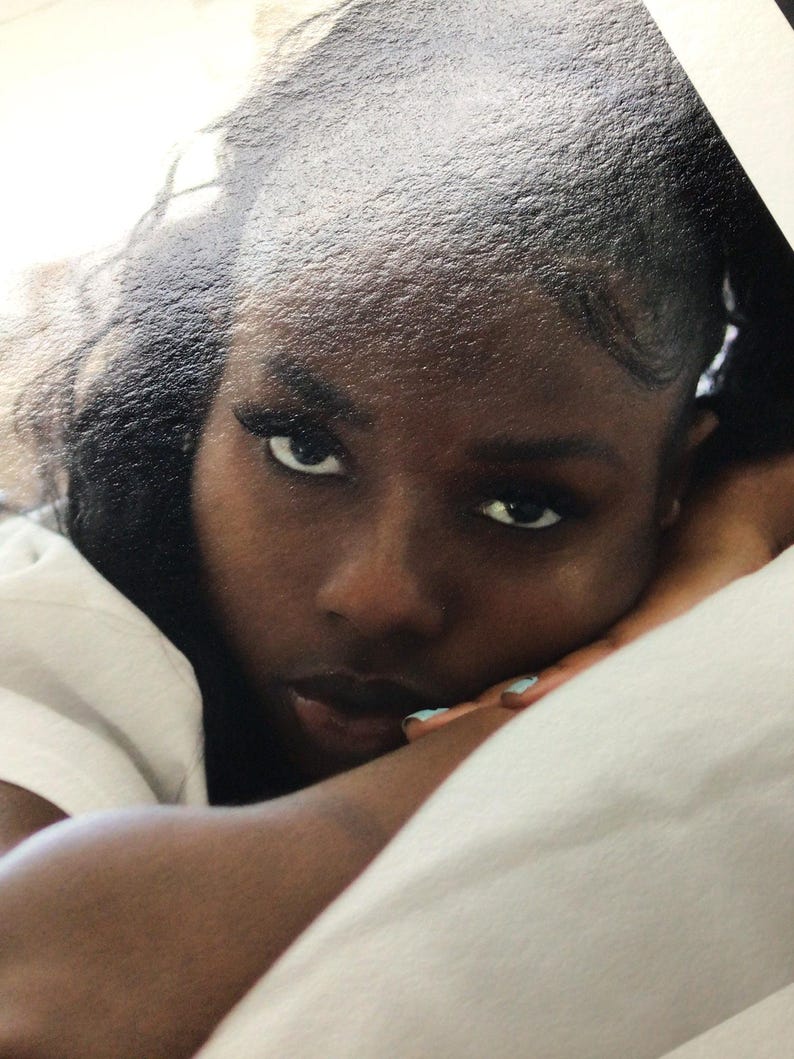 May include: A portrait of a person with dark skin, dark hair styled back, and dark eyes looking directly forward. The person is resting their chin on their arms, which are positioned on a white surface. The person wears light blue nail polish.