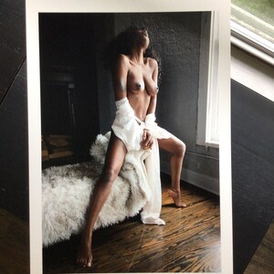 May include: A photographic print featuring a person with dark skin and long dark hair, posed in a seated position. The person is wearing a white, sheer, flowing garment draped around the waist and hips, set against a dark, moody interior.