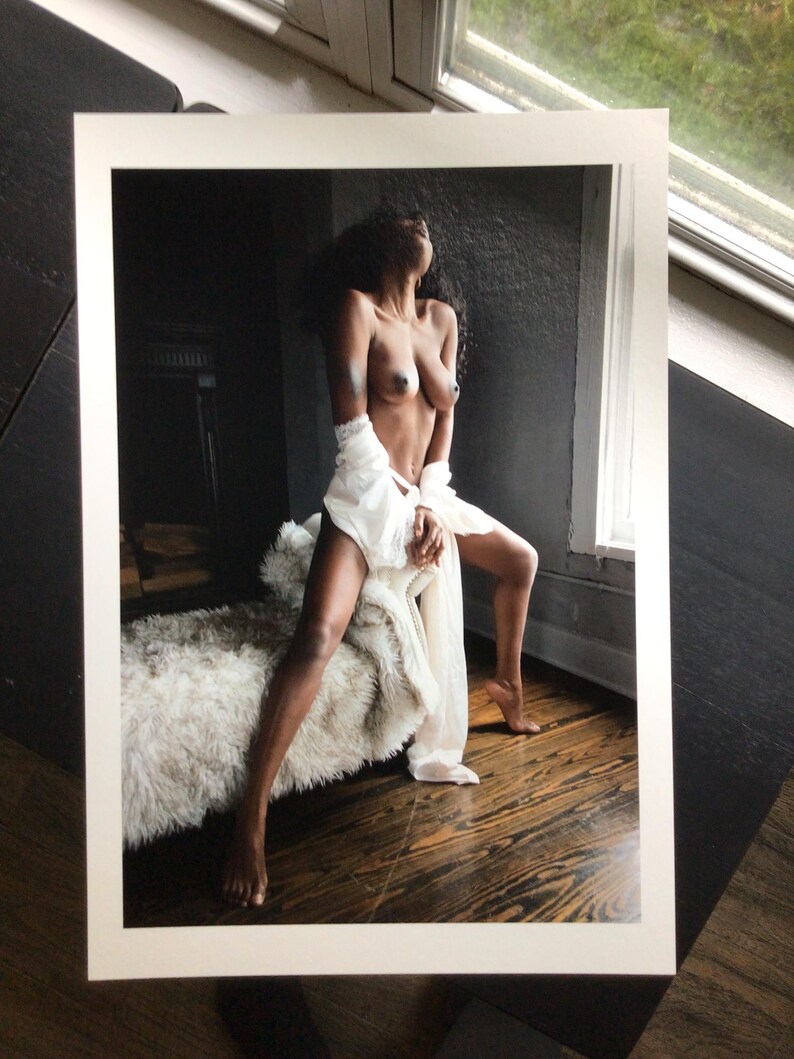 May include: A photographic print featuring a person with dark skin and long dark hair, posed in a seated position. The person is wearing a white, sheer, flowing garment draped around the waist and hips, set against a dark, moody interior.