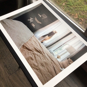 May include: A framed print featuring a bedroom scene with a bed covered in a tan comforter and a textured, light-colored throw. The print is set against a window with sheer curtains, and the frame is black.