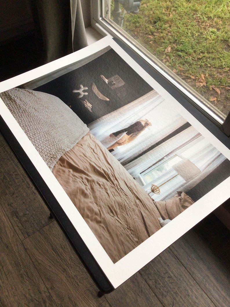 May include: A framed print featuring a bedroom scene with a bed covered in a tan comforter and a textured, light-colored throw. The print is set against a window with sheer curtains, and the frame is black.