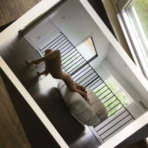 May include: A rectangular wall mirror with a thick, clean white frame. The mirror surface reflects a room interior featuring a dark wooden floor, a white tufted sofa, and a black metal staircase railing, capturing a clear view of the indoor space.
