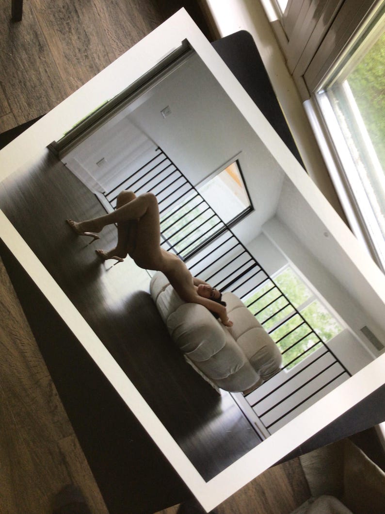 May include: A rectangular wall mirror with a thick, clean white frame. The mirror surface reflects a room interior featuring a dark wooden floor, a white tufted sofa, and a black metal staircase railing, capturing a clear view of the indoor space.