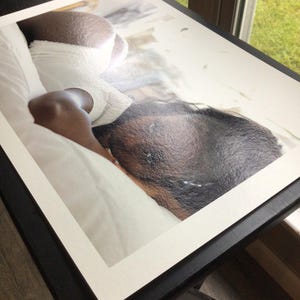 May include: A rectangular photographic print featuring a high-angle view of a person with dark skin lying on white bedding. The person is wearing white textured clothing and is positioned on their side, with their head resting on the white fabric.