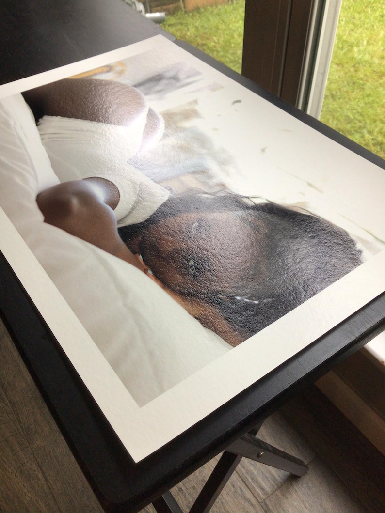 May include: A rectangular photographic print featuring a high-angle view of a person with dark skin lying on white bedding. The person is wearing white textured clothing and is positioned on their side, with their head resting on the white fabric.