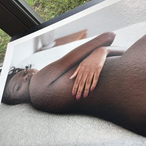 May include: A print of a person lying down, with dark skin and dark hair. The person's arm is bent, and their hand is resting on their body. The person has pink nail polish. The print is on a white surface.