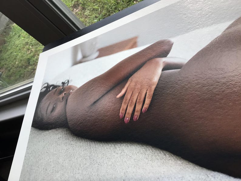 May include: A print of a person lying down, with dark skin and dark hair. The person's arm is bent, and their hand is resting on their body. The person has pink nail polish. The print is on a white surface.