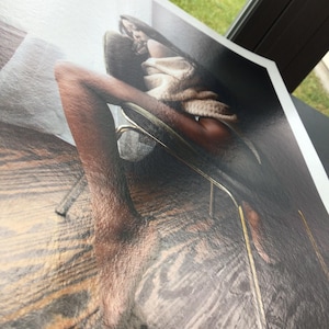 May include: A print of a person in a chair, with a neutral-toned sweater. The image has a glossy finish and is angled, showing the edge of the print. The background is blurred, with a view of a window and greenery.