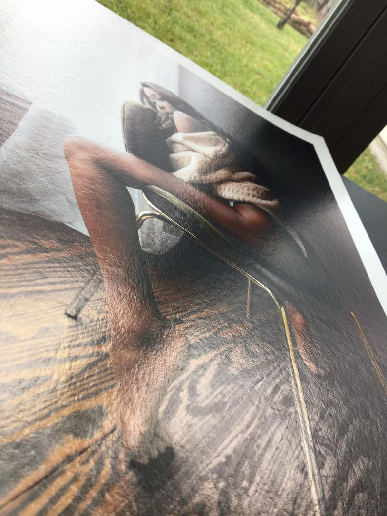 May include: A print of a person in a chair, with a neutral-toned sweater. The image has a glossy finish and is angled, showing the edge of the print. The background is blurred, with a view of a window and greenery.