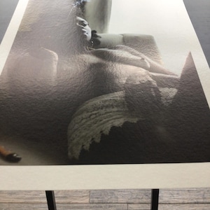 May include: Monochrome art print depicting a person reclining. The image has a textured finish, showing a figure with visible facial features and a dark backdrop. The print is displayed on a black stand.