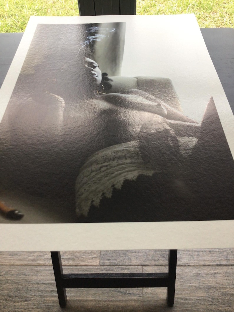 May include: Monochrome art print depicting a person reclining. The image has a textured finish, showing a figure with visible facial features and a dark backdrop. The print is displayed on a black stand.
