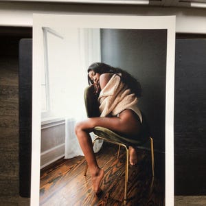 May include: A print of a person sitting on a green velvet chair with gold legs. The person is wearing a beige sweater and white underwear. The background includes a window with white curtains and dark wood flooring.