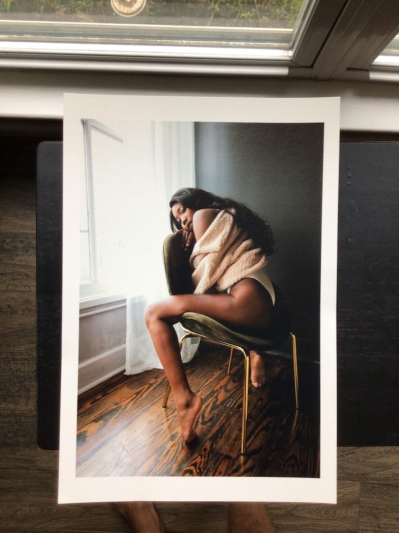 May include: A print of a person sitting on a green velvet chair with gold legs. The person is wearing a beige sweater and white underwear. The background includes a window with white curtains and dark wood flooring.