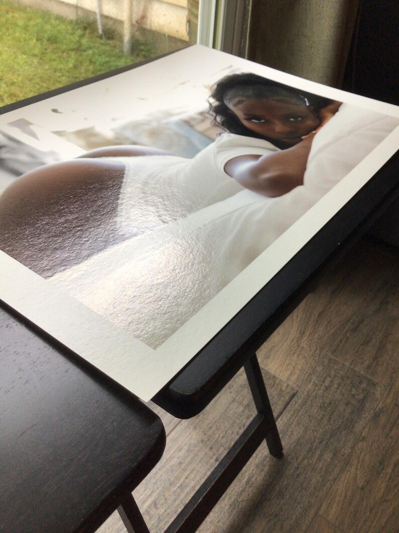 May include: A rectangular photographic print featuring a person with dark skin wearing a white bodysuit, lying down and looking toward the camera. The print has a white border and a glossy finish, displaying a high-contrast composition of light and shadow.