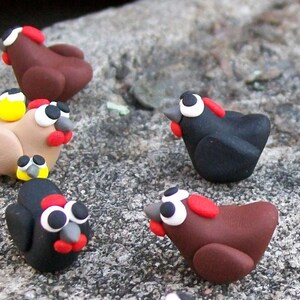 May include: A collection of handmade clay chickens in various colors, including brown, black, and beige. Each chicken has a red comb and wattle, and black eyes. The chickens are arranged on a textured surface.