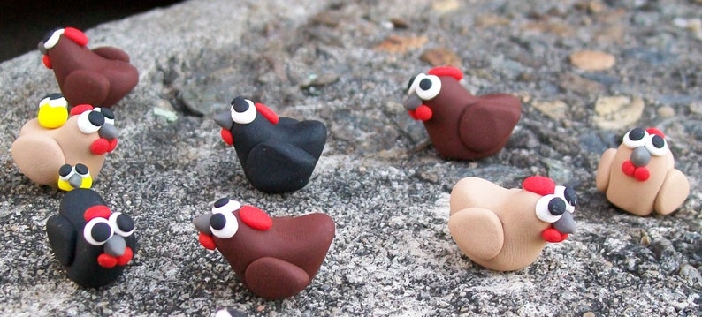 May include: A collection of handmade clay chickens in various colors, including brown, black, and beige. Each chicken has a red comb and wattle, and black eyes. The chickens are arranged on a textured surface.