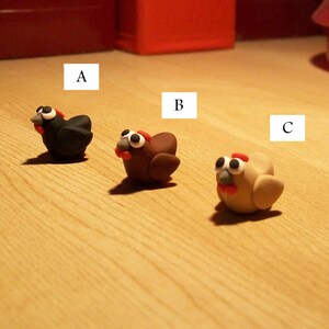 May include: Three small clay figurines of chickens. The first chicken is black, the second is brown, and the third is beige. The chickens are labeled A, B, and C.
