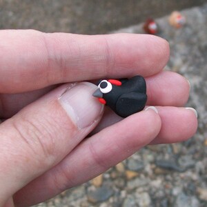 May include: A small black bird figurine with red accents and white eyes. The figurine is made of clay and is being held in a person's hand.
