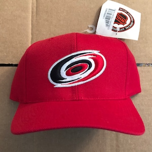 May include: Red baseball cap with a white, black, and red Carolina Hurricanes logo embroidered on the front. The cap has a curved brim and an adjustable strap.