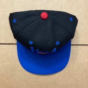Puede incluir: Una gorra de b&eacute;isbol negra y azul con un bot&oacute;n rojo en la parte superior. La parte delantera de la gorra tiene la palabra "NBA" bordada en azul.