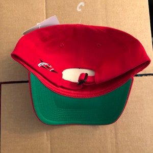 May include: Red baseball cap with a green underside. The cap has a black adjustable strap and a white logo on the back.