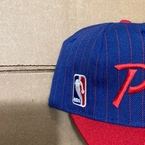 Op de afbeelding: Een blauwe en rode baseballpet met een rode "P" op de voorkant en een NBA-logo aan de zijkant. De pet heeft een rode klep en een pinstripe-patroon.