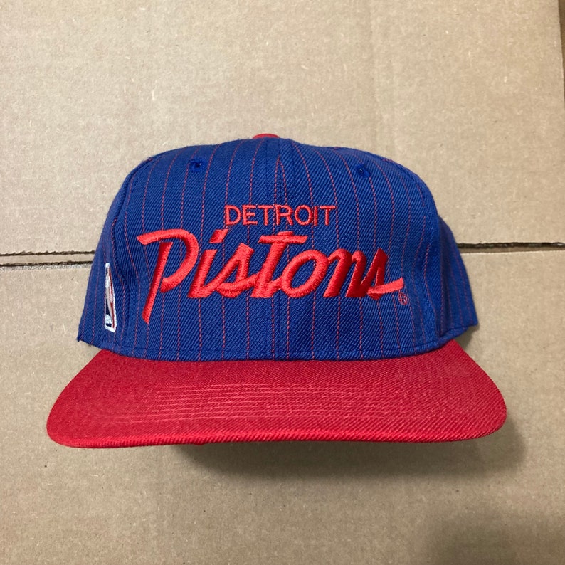 May include: A blue and red baseball cap with a white pinstripe pattern. The cap has the text "DETROIT" and "Pistons" embroidered in red on the front.
