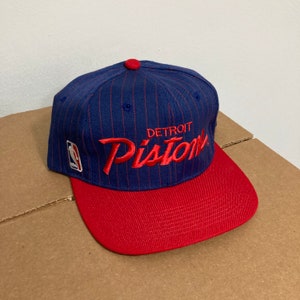 Op de afbeelding: Een blauwe en rode baseballpet met een wit NBA-logo en de tekst "DETROIT PISTONS" in rode borduurwerk.