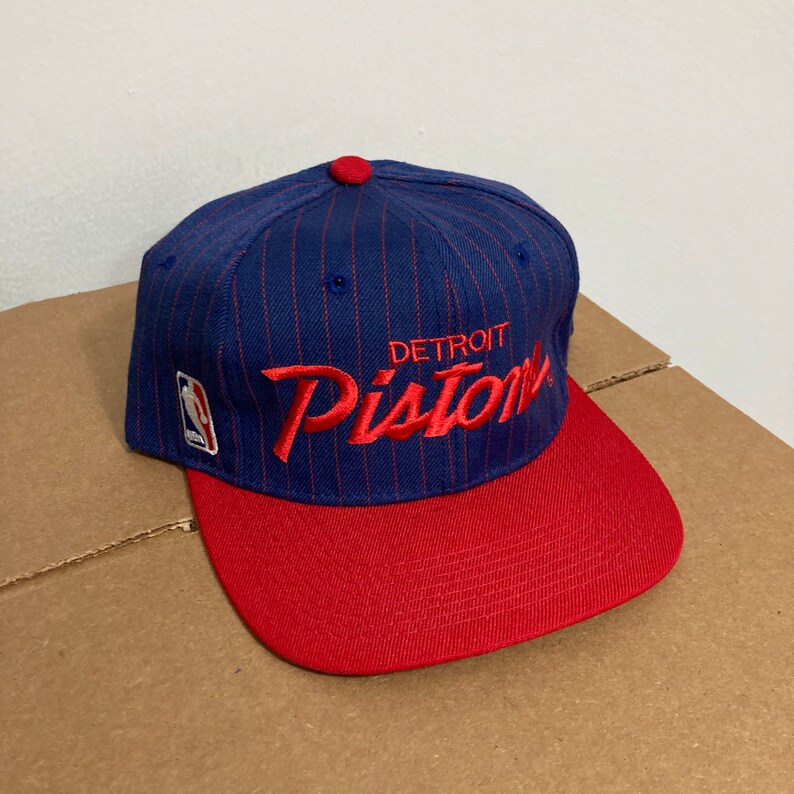 Op de afbeelding: Een blauwe en rode baseballpet met een wit NBA-logo en de tekst "DETROIT PISTONS" in rode borduurwerk.