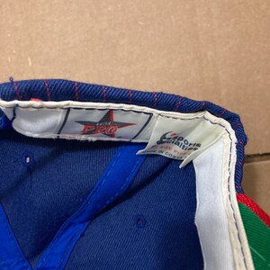 Op de afbeelding: Een blauwe en witte baseballpet met een rode en groene klep. De pet heeft een witte ster met de tekst "The Pro" erin. De achterkant van de pet heeft een wit label met de tekst "Sports Specialties" en "Size Fits All" in zwarte tekst. Het label vermeldt ook "Made in Costa Rica".