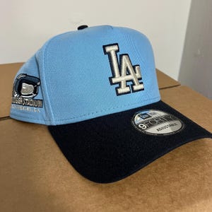 May include: A light blue and black adjustable baseball cap. The front of the cap features the white and gray embroidered letters "LA". The side of the cap has a patch that says "DODGER STADIUM LOS ANGELES, CA".