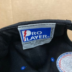 Puede incluir: Una gorra de b&eacute;isbol negra con un logotipo azul y blanco. La etiqueta en el interior de la gorra dice "Pro Layer" y "Hecho en Bangladesh 80% Poli&eacute;ster 20% Lana".