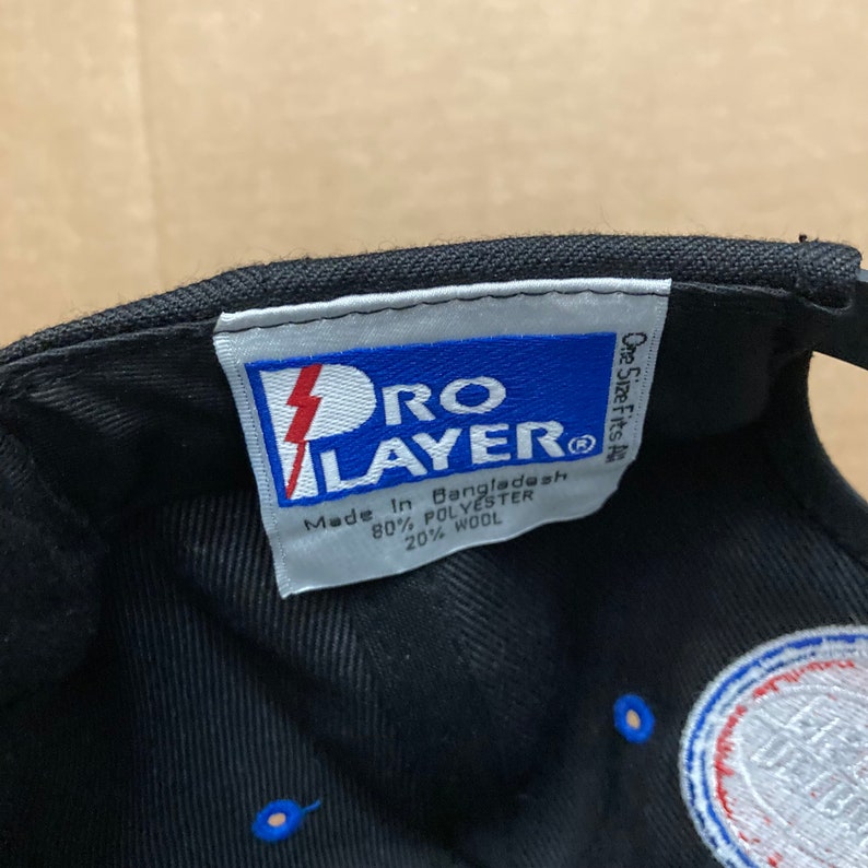 Puede incluir: Una gorra de b&eacute;isbol negra con un logotipo azul y blanco. La etiqueta en el interior de la gorra dice "Pro Layer" y "Hecho en Bangladesh 80% Poli&eacute;ster 20% Lana".