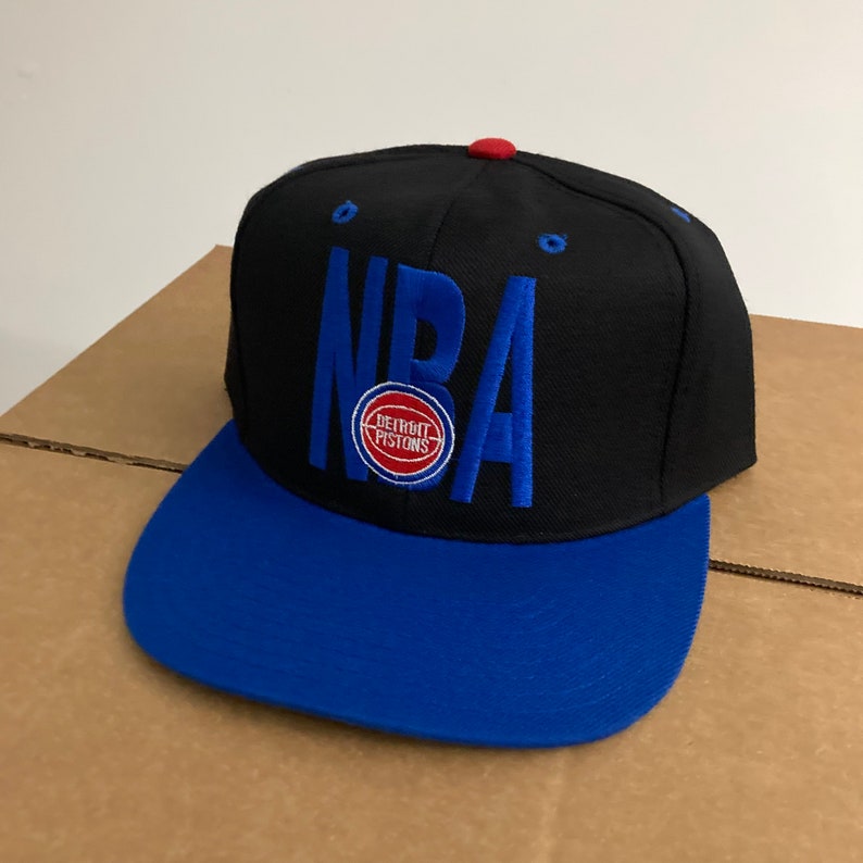 Puede incluir: Gorra de b&eacute;isbol negra y azul con la palabra "NBA" bordada en azul en la parte delantera. La gorra tambi&eacute;n presenta un logotipo de los Detroit Pistons en rojo y azul.