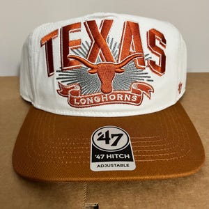 Peut inclure: Casquette de baseball blanche et marron avec le mot "TEXAS" brodé en orange. La casquette présente un logo Longhorns et une visière marron. L'étiquette indique "47 Hitch Adjustable".