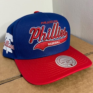 May include: A blue and red Philadelphia Phillies baseball cap. The cap features the team name in red script with a white outline, and the word "BASEBALL" in red. The brim is red, and the cap has a flat bill.
