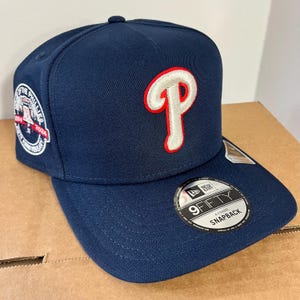 May include: A navy blue snapback baseball cap featuring the Philadelphia Phillies logo in white with a red outline. The cap has a flat brim and a 9FIFTY sticker. The side of the cap displays a team anniversary patch.