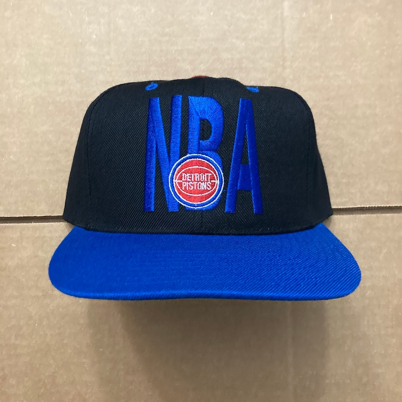 May include: Black and blue baseball cap with the word "NBA" embroidered on the front. The cap also features a red and blue Detroit Pistons logo.