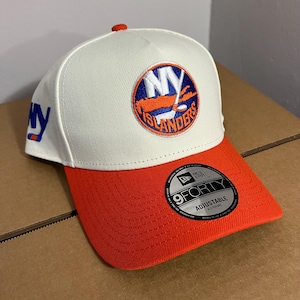 May include: A white and orange New York Islanders baseball cap. The cap features the team's logo on the front and the letters "NY" on the side. The brim is orange, and the cap is adjustable.