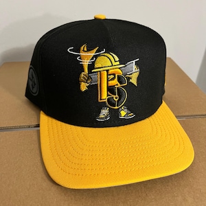 May include: A black baseball cap with a yellow brim and a yellow button on top. The front features an embroidered graphic of a stylised letter B wearing a yellow hard hat, holding a wrench and a steel beam, with small trainers on its feet.