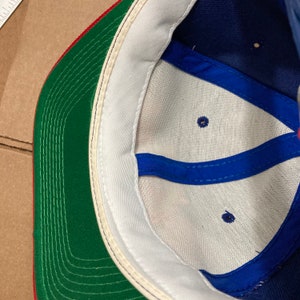 Op de afbeelding: De binnenkant van een baseballpet met een witte kroon, een groene klep en een blauwe onderkant. De pet heeft een blauw bandje met twee kleine gaatjes.