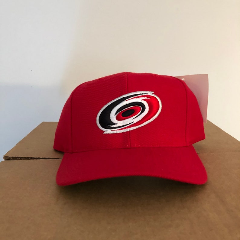 May include: Red baseball cap with a white, black, and red Carolina Hurricanes logo embroidered on the front.
