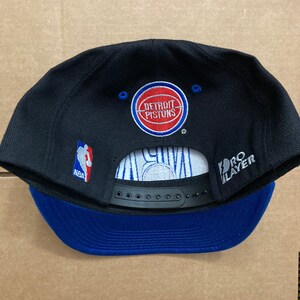Puede incluir: Gorra de b&eacute;isbol negra y azul con un logotipo de Detroit Pistons blanco y azul en la parte delantera. La gorra tiene una correa negra con un logotipo "NBA" blanco y una correa negra con un logotipo "Pro Player" blanco en la parte posterior.