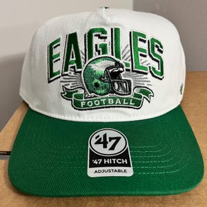 May include: White and green baseball cap with the word "EAGLES" embroidered in green. The cap features a green football helmet graphic and the word "FOOTBALL" on a banner. The brim is green, and the tag reads "'47 Hitch Adjustable".