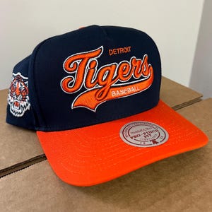 May include: A navy and orange baseball cap with the Detroit Tigers logo in orange script. The cap has a flat peak and a tiger head patch on the side. The cap is a Pro-Fit structured style.