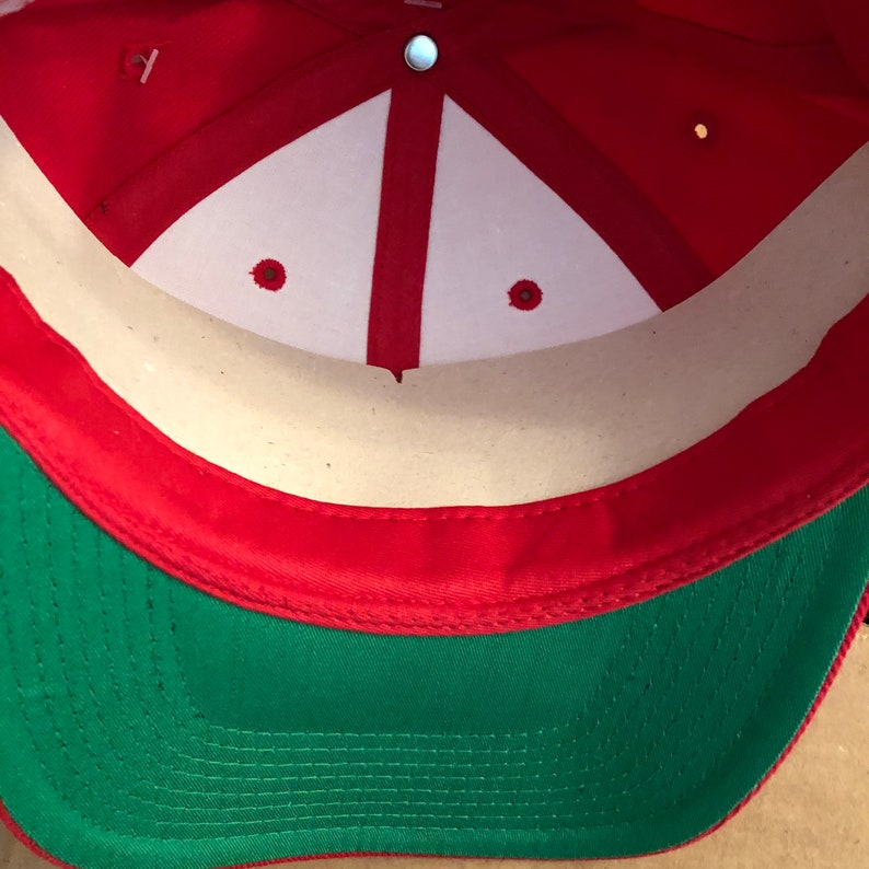 May include: The inside of a red and green baseball cap. The cap has a white sweatband and a red brim. The inside of the cap is made of a beige fabric.
