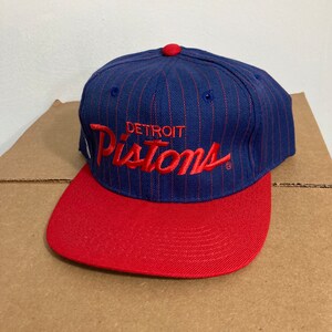 Op de afbeelding: Een blauwe en rode baseballpet met een wit pinstripe patroon. De pet heeft de woorden "DETROIT PISTONS" in rood geborduurd op de voorkant.